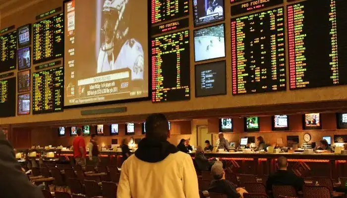 A sports betting venue displaying large screens and betting odds boards, with patrons engaged in various activities.