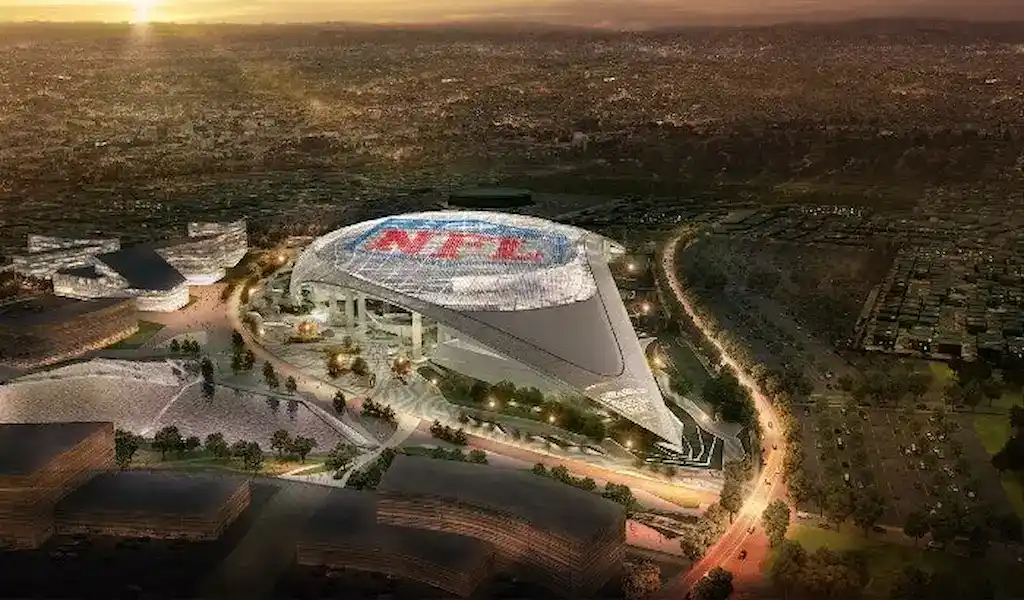 An aerial view of a modern NFL stadium at sunset, featuring a large NFL logo on the roof, surrounded by surrounding buildings, parking areas, and a cityscape in the background.