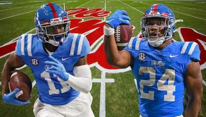 Two Ole Miss football players in blue uniforms pose with footballs on a field featuring the Ole Miss logo in the background.