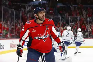 A Washington Capitals player reacts with excitement on the ice, with Toronto Maple Leafs players blurred in the background.