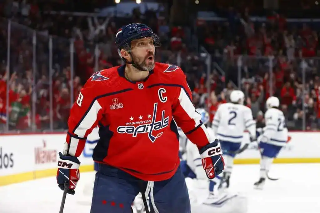 A Washington Capitals player reacts with excitement on the ice, with Toronto Maple Leafs players blurred in the background.