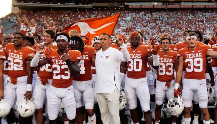 Texas Longhorns football players and coach standing together with fans and a team flag in the background.