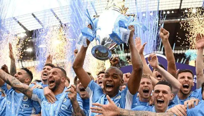Gambling News 9 Manchester City players jubilantly celebrate as their captain lifts the Premier League trophy with confetti and fireworks in the background.