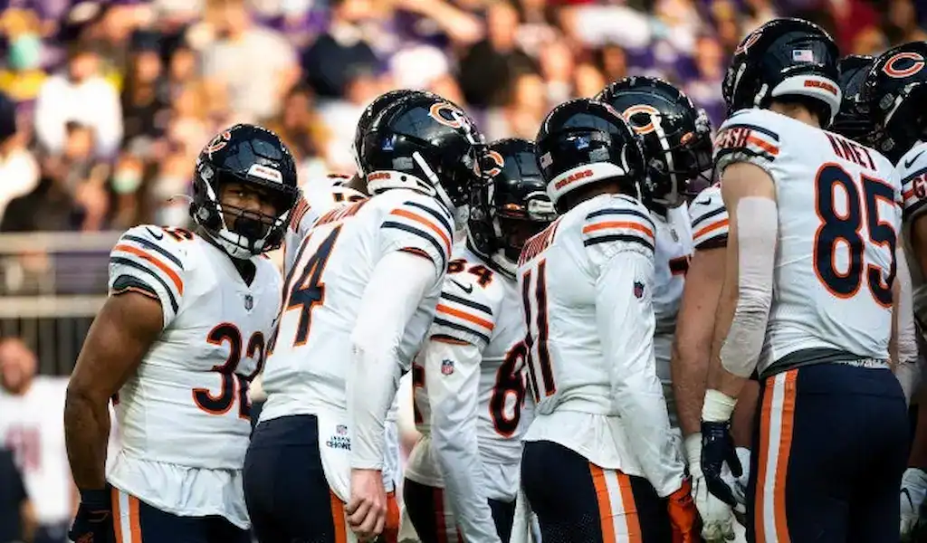 Chicago Bears players huddle on the field during an NFL game wearing white jerseys with navy helmets and orange accents.