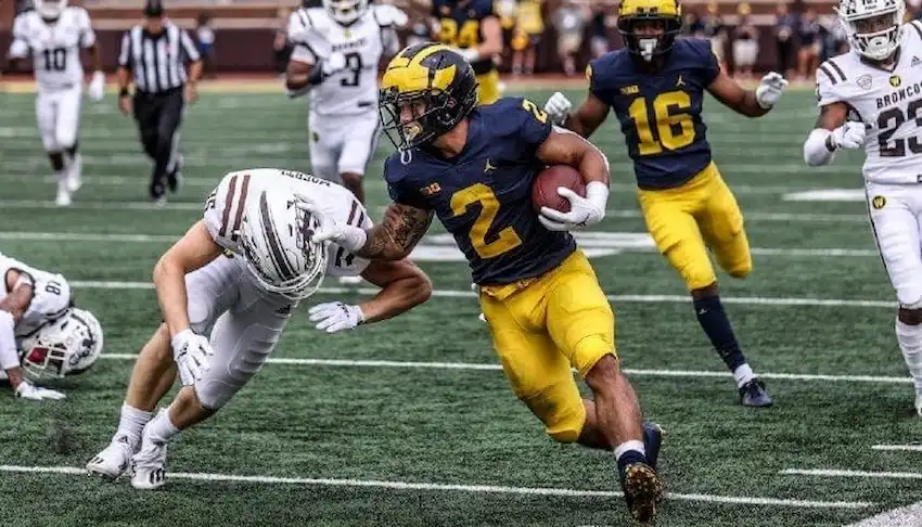 A college football player runs with the ball while stiff-arming a defender during a game.