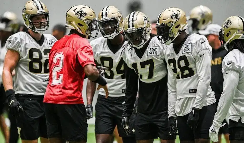 New Orleans Saints players huddle during practice in helmets and uniforms as they prepare for the upcoming NFL season.
