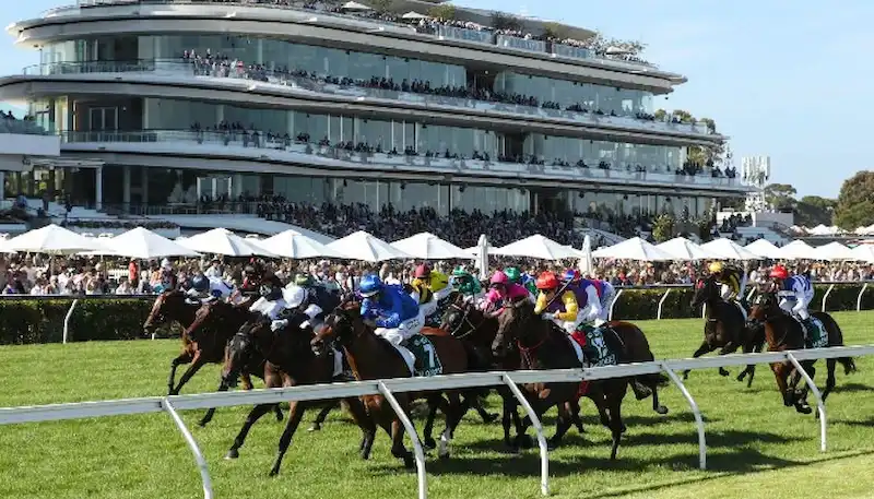 A group of jockeys on horseback race down the turf track.