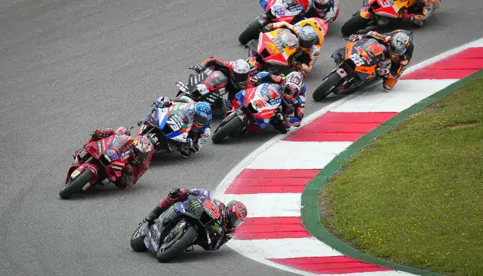 MotoGP riders leaning into a sharp corner during a high-speed race, closely packed together as they battle for position on the track.