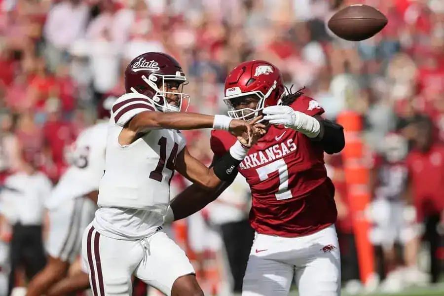 A football player in an Arkansas Razorbacks red jersey pressures the opposing quarterback as the ball is released.
