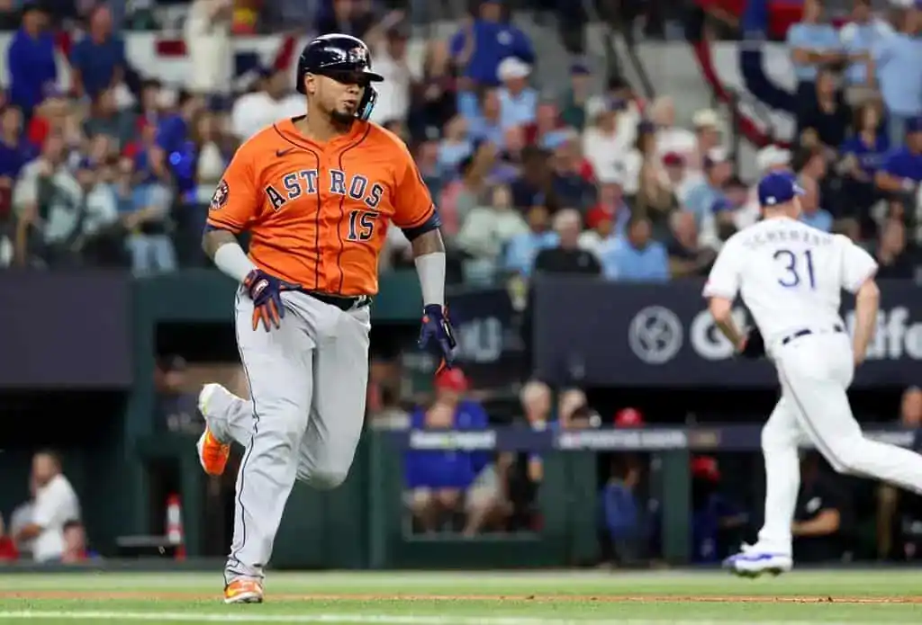 An Astros player in an orange jersey sprints towards home plate as a pitcher in white prepares to throw. Fans watch excitedly in the background.