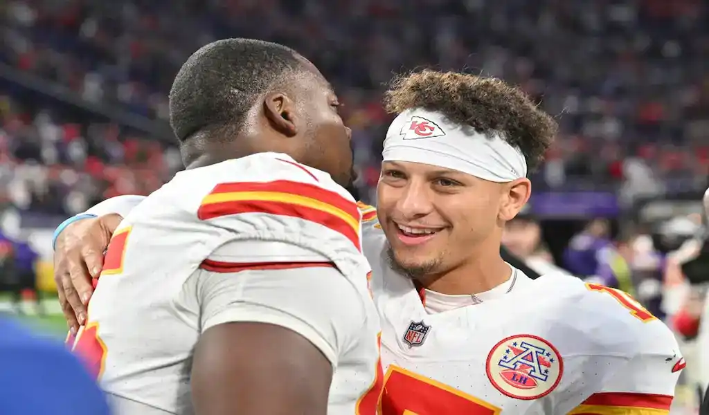 2 Kansas City Chiefs players celebrate together after a game, with Patrick Mahomes smiling and hugging a teammate after a successful play on the field.