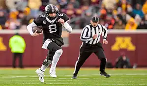 Minnesota Golden Gophers quarterback running with the ball during a college football game as a referee follows the play.