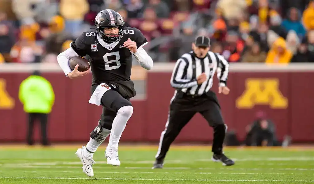 Minnesota Golden Gophers quarterback running with the ball during a college football game as a referee follows the play.