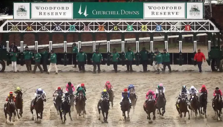 Horses burst out of the starting gates at Churchill Downs during a race on a muddy track.