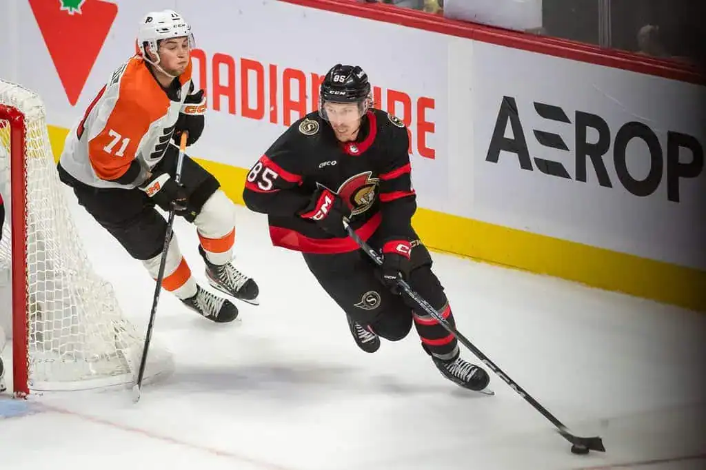 A hockey player in a black jersey races past an opponent in orange and black, with a goal net visible in the background.