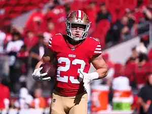 San Francisco 49ers player wearing jersey number 23 runs with the football during a game, with the crowd blurred in the background.