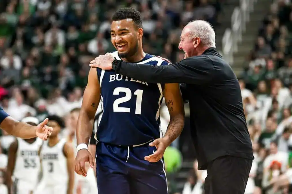 A basketball player in a Butler uniform receives guidance from a coach during an intense game, with a cheering crowd in the background.