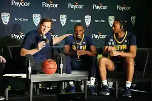 California Golden Bears coach and players smiling and speaking at a PAC-12 press conference with microphones and basketballs on stage.