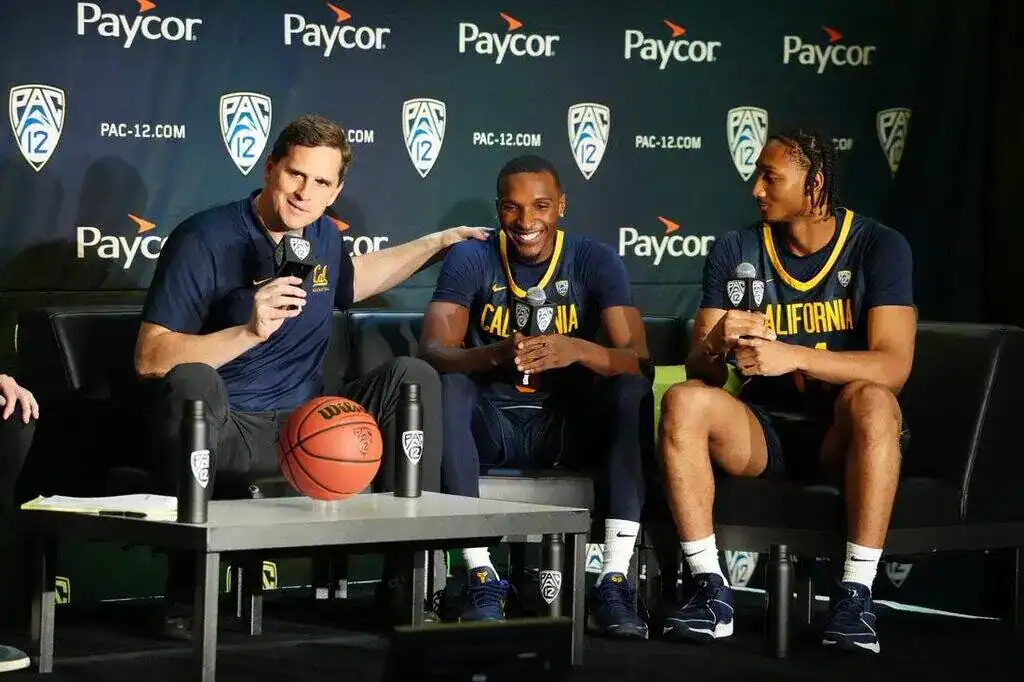 California Golden Bears coach and players smiling and speaking at a PAC-12 press conference with microphones and basketballs on stage.