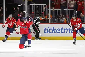 Washington Capitals player celebrating a goal on one knee while teammates skate in to join.
