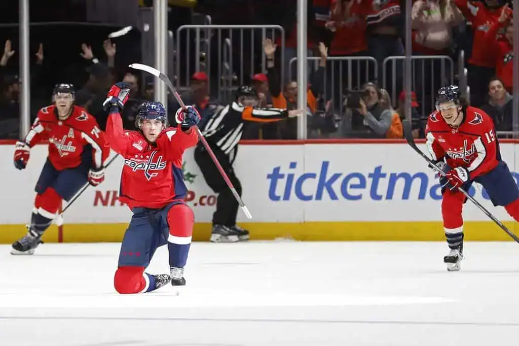 Washington Capitals player celebrating a goal on one knee while teammates skate in to join.