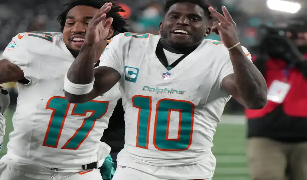 7 Miami Dolphins players celebrate on the field wearing white uniforms with teal and orange accents during an NFL game.