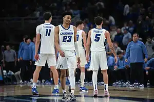 A group of basketball players in white jerseys with blue accents stand on the court, preparing for a game, with fans in the background.