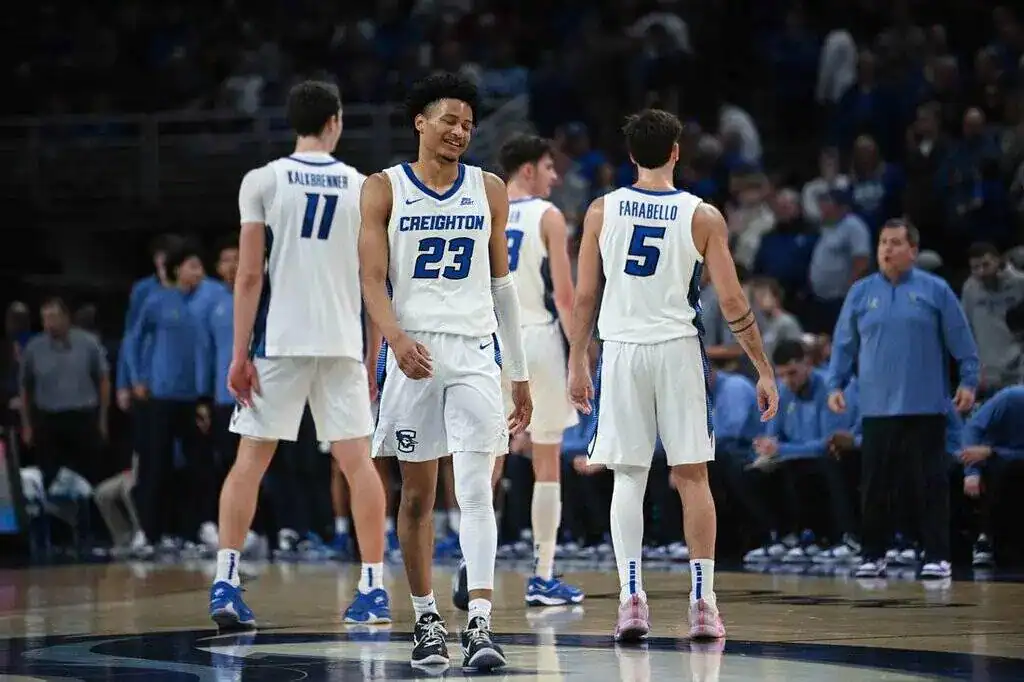 A group of basketball players in white jerseys with blue accents stand on the court, preparing for a game, with fans in the background.