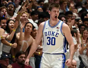 Duke basketball player in a white uniform shouting in celebration as fans cheer in the background.