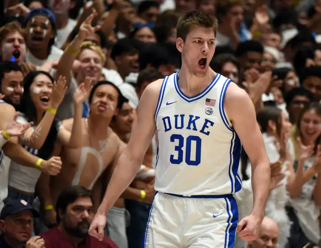 Duke basketball player in a white uniform shouting in celebration as fans cheer in the background.