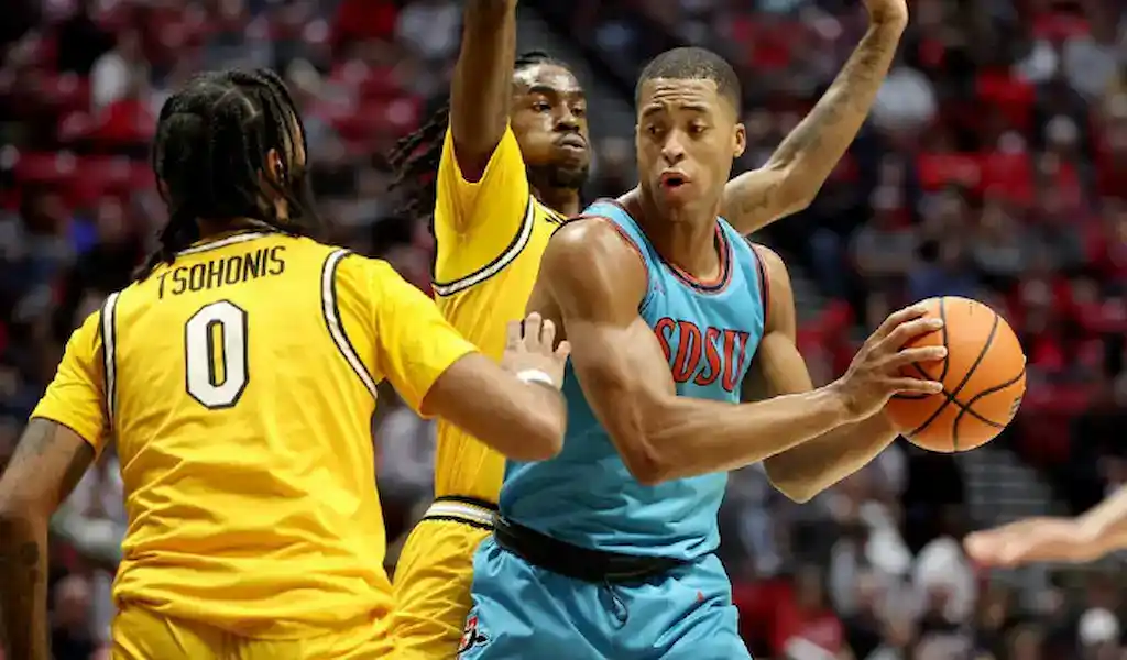 Two NCAA basketball players defend against a South Dakota State player during a game, highlighting team strategy and totals betting.