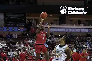 A basketball player in a red Houston jersey shoots the ball, while a competitor in a yellow Towson jersey defends. Spectators fill the stands.