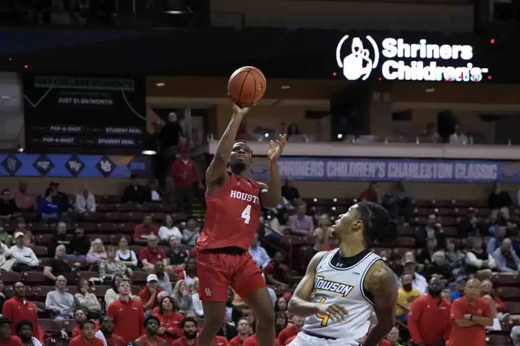 A basketball player in a red Houston jersey shoots the ball, while a competitor in a yellow Towson jersey defends. Spectators fill the stands.