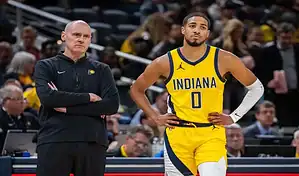 An Indiana basketball player in a yellow and blue uniform with the number 0 standing on the court, next to a coach with arms crossed, with a blurred crowd in the background.