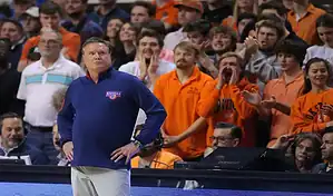 Kansas Jayhawks head coach stands courtside during a college basketball game as fans in the background cheer and react.