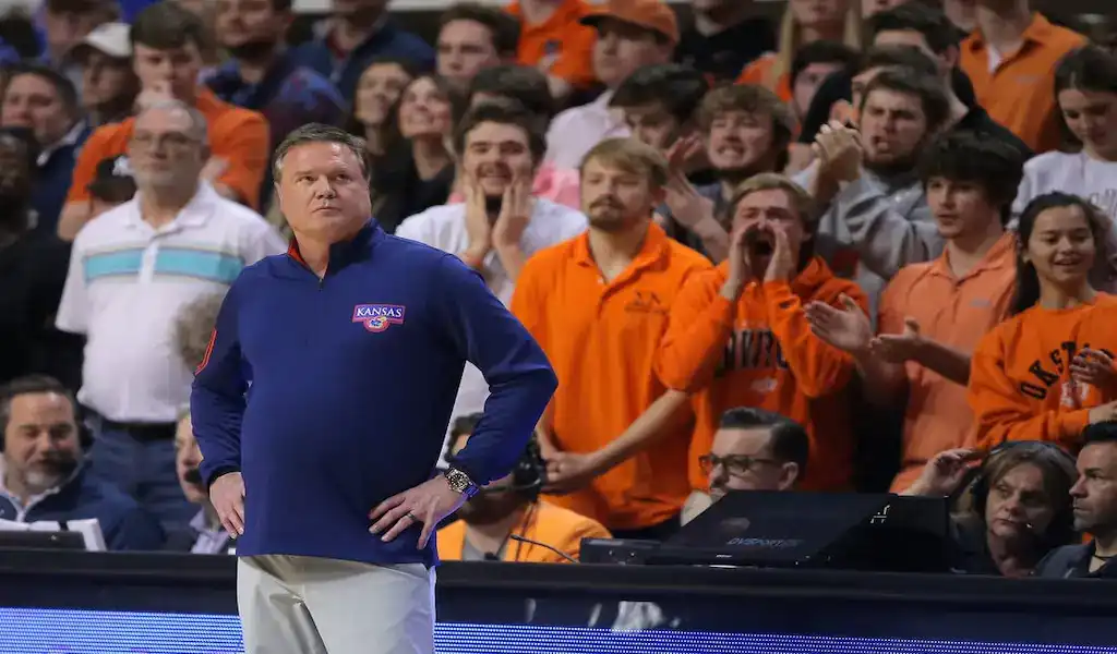 Kansas Jayhawks head coach stands courtside during a college basketball game as fans in the background cheer and react.