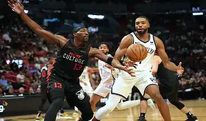 A Brooklyn Nets player dribbling the ball during an intense game, guarded by a Miami Heat player in a black and red uniform with the number 13, with a blurred crowd in the background.
