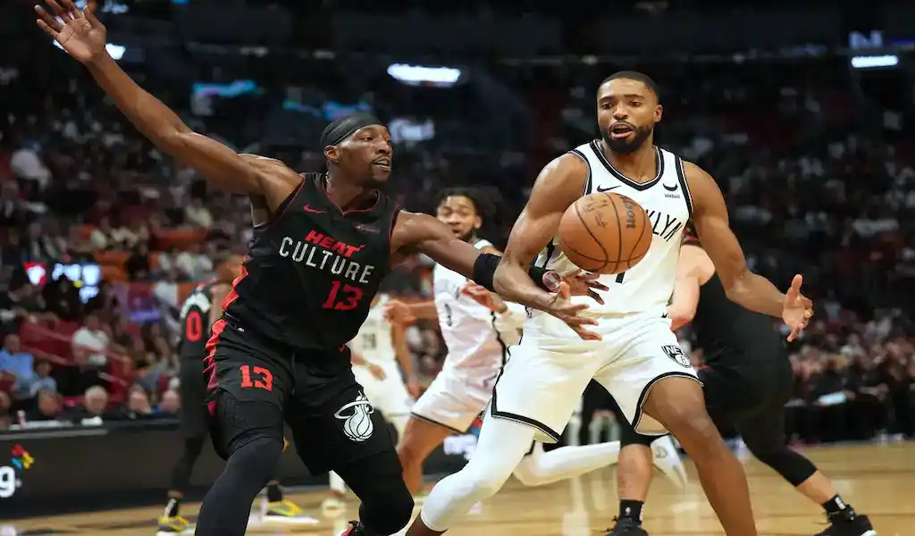 2 A Brooklyn Nets player dribbling the ball during an intense game, guarded by a Miami Heat player in a black and red uniform with the number 13, with a blurred crowd in the background.