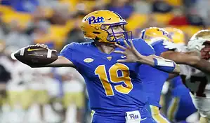 A Pitt quarterback in a blue and yellow uniform with the number 19, throwing a football during a game, with a blurred crowd and opposing player in the background.