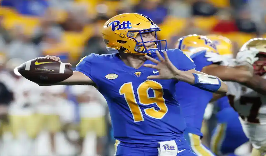 A Pitt quarterback in a blue and yellow uniform with the number 19, throwing a football during a game, with a blurred crowd and opposing player in the background.