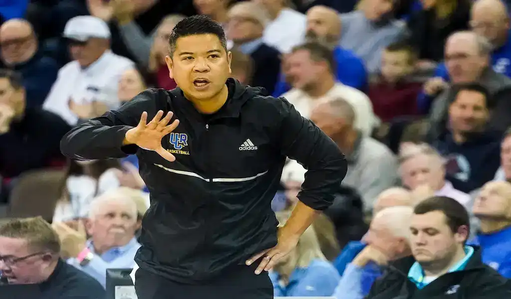 College basketball coach giving instructions from the sideline during a game, with fans watching in the background.