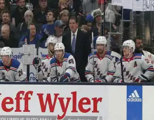 New York Rangers players and coaching staff watch the game intently from the bench during a match against the Carolina Hurricanes, with fans visible behind the glass.