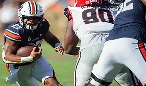 An Arkansas Razorbacks player in a red and white uniform with number 7 running with the ball during a game against an Auburn Tigers player in an orange and blue uniform with number 9, with a blurred crowd in the background, dated November 11, 2023.