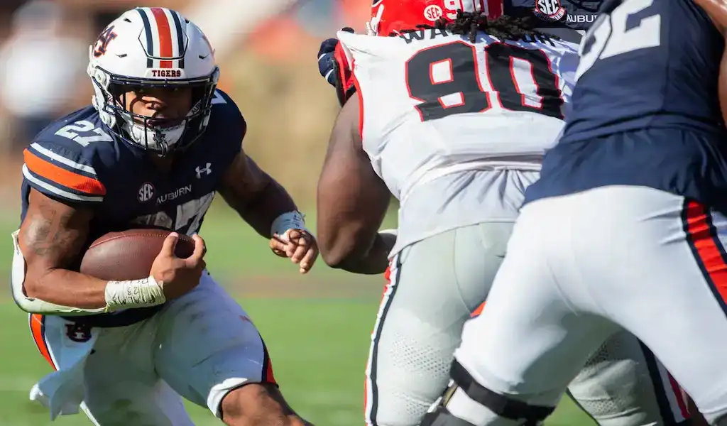 An Arkansas Razorbacks player in a red and white uniform with number 7 running with the ball during a game against an Auburn Tigers player in an orange and blue uniform with number 9, with a blurred crowd in the background, dated November 11, 2023.