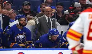 A Buffalo Sabres coach and players, including one with number 53, on the bench during a game against the Minnesota Wild, with a blurred crowd and opposing player in the foreground, dated November 10, 2023.