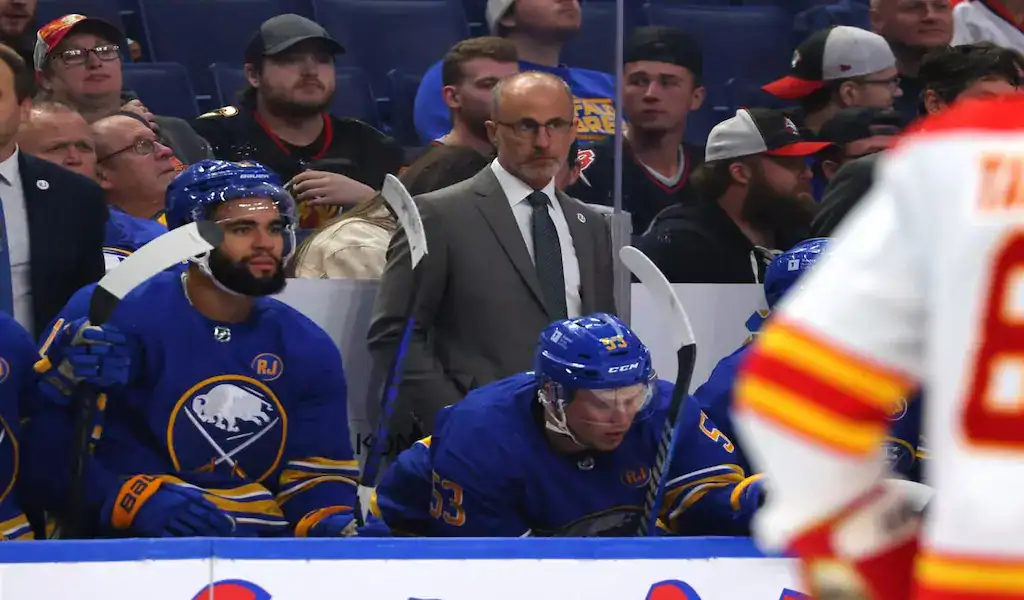 NHL 9 A Buffalo Sabres coach and players, including one with number 53, on the bench during a game against the Minnesota Wild, with a blurred crowd and opposing player in the foreground, dated November 10, 2023.