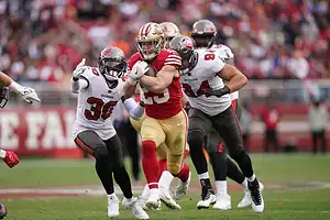 49ers running back Christian McCaffrey runs with the ball, pursued by Buccaneers defenders.