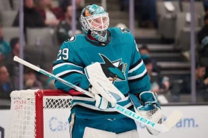 A San Jose Sharks goalie in teal gear and a decorated helmet stands in front of the net during a game.