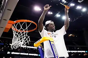 A player celebrates on a ladder, holding scissors high above the basketball hoop, ready to cut down the net after a win.