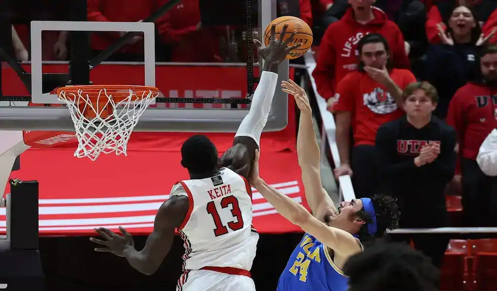 A St. John’s Red Storm player in a white uniform with number 13 attempting a shot near the basket, defended by a Utah Utes player in a blue and yellow uniform with number 24, with a crowd in red in the background, dated November 19, 2023.